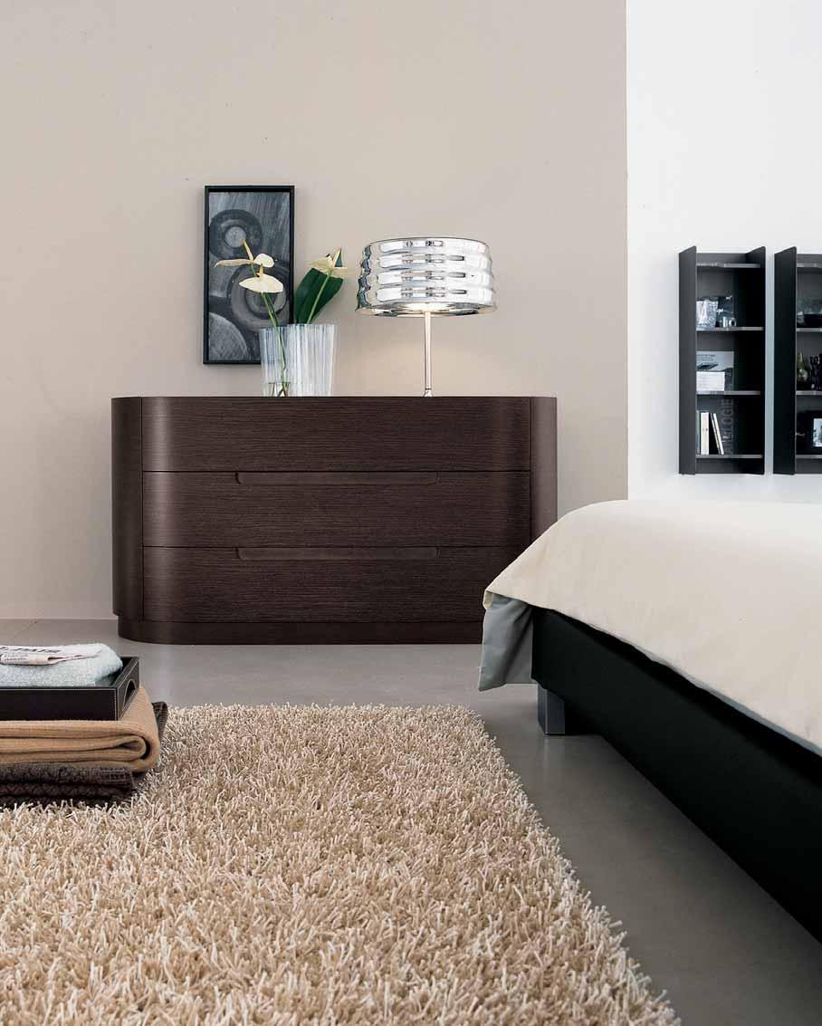 SINTESI BED UNIT IN COFFEE-COLOURED OAK. BEDSIDE TABLE Ø 54 H 38,6. CHEST DRAWERS W 140 D 55 H 73,8. TECH WALL UNITS IN ALUMINIUM TITANIUM FINISH W 35 D 28 H 114,2 AND GLOSS WHITE LACQUERED SHELVES.