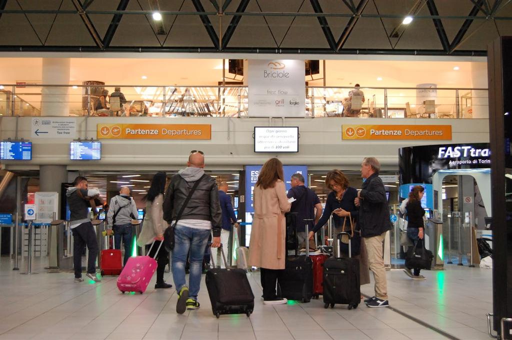 Nel terminal Area Partenze Tante persone con le valigie, cartelloni pubblicitari, schermi con i voli in partenza. Voci, annunci preceduti da un segnale, musica, rumore delle valigie.