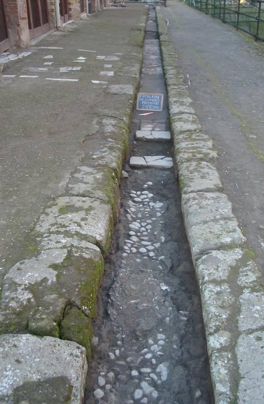 10: La canaletta rivestita di ciottoli, il marciapiede del lato Sud del Decumano Massimo ed il fusto di una colonna dipinto in rosso visibile nel cunicolo. Fig.