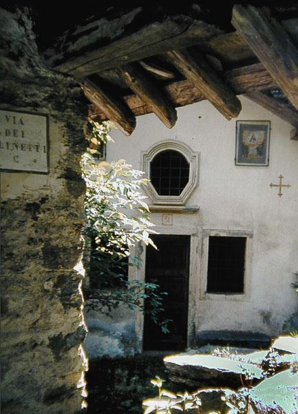 Facciata e abside della cappella della Trinità. Sul muro del mulino antistante la chiesetta si vede l'antica targa viaria che indica la Via dei Molinetti, sopraelevata rispetto alla piazzuola.